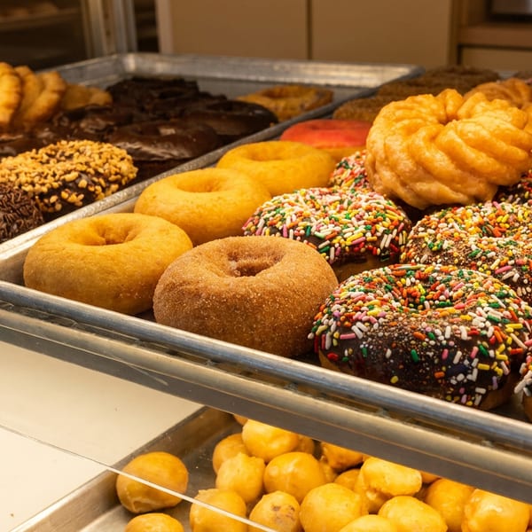 Trays display assorted glazed, powdered, chocolate-covered, and sprinkle-covered donuts in a bakery case.